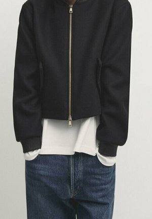 Person trägt eine schwarze Zip-up-Jacke über einem weißen Shirt mit blauen Jeans, Hände in den Taschen, steht vor einem schlichten Hintergrund.