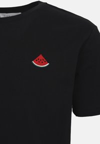 Μαύρο μπλουζάκι με κοντά μανίκια και μικρό κεντημένο κόκκινο κομμάτι καρπούζι με μαύρα σπόρια στην αριστερή πλευρά του στήθους.