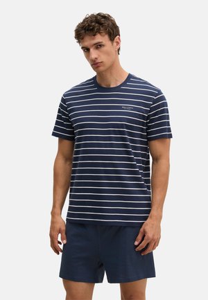 Jonge man die een marineblauw-wit gestreept T-shirt met korte mouwen en effen marineblauwe short draagt en voor een effen achtergrond staat.