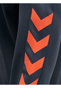 Pantalon de sport bleu marine avec motif chevron rouge sur le côté, tissu texturé et coupe fuselée pour un look ajusté.