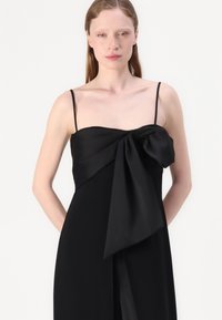 Robe noire avec des bretelles spaghetti, ornée d'un grand nœud à l'avant, fabriquée en tissu lisse et léger, avec une silhouette fluide.