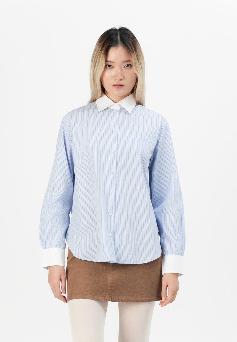 Light blue button-up shirt with thin vertical stripes, white collar, and cuffs. Worn with a brown skirt. Smooth, textured fabric.