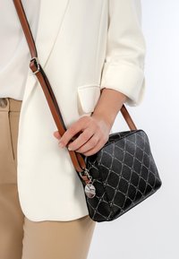 Person wearing beige pants and white blazer holding a black patterned leather crossbody bag with brown strap and silver charm.