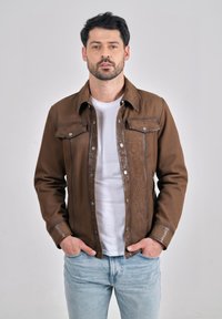 Mand med mørkt hår og skæg iført brun jakke over hvid skjorte og lyseblå jeans, stående med hænderne i lommerne mod en ensfarvet baggrund.