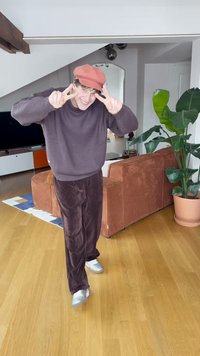 Személy bordó kötött pulóverben, sötétbarna kordbársony nadrágban és világos narancssárga sapkában. Beltéren áll, háttérben növény és kanapé látható.