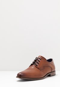 Chaussure habillée en cuir marron avec un laçage, texture lisse, bout rond et un talon fin. Fines surpiqûres sur les côtés.