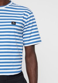 Muž v modro-bílém horizontálně pruhovaném tričku s krátkým rukávem a malým černobílým logem na hrudi, doplněný černými kalhotami.