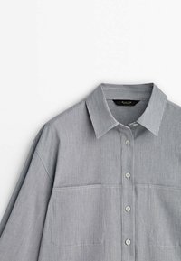 Chemise gris clair à manches longues en tissu texturé, avec un col évasé, deux poches avant et des boutons blancs à rayures verticales.