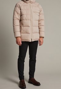 Beige puffjacka med quiltad textur, framdragkedja och huva. Buren över svarta byxor och mörkbruna stövlar med gummisula.