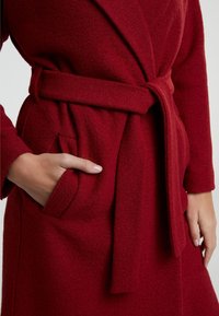 Manteau rouge texturé avec une taille ceinturée, comprenant un col châle et des poches latérales. Gros plan sur une main dans la poche, mettant en valeur les détails du tissu.
