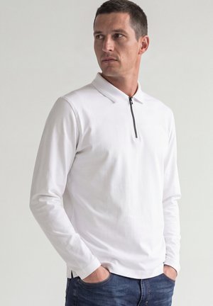 Homme portant un polo blanc à manches longues avec une fermeture éclair devant, mains dans les poches, regardant sur le côté sur un fond uni.
