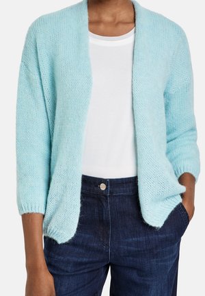 Personne portant un cardigan bleu clair ouvert sur un t-shirt blanc et un jean bleu foncé, une main dans la poche.