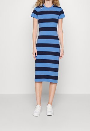Kobieta w niebiesko-granatowej sukience w poziome paski do kolan, z krótkimi rękawami i białych trampkach, stojąca na jednolitym tle.