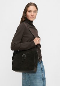 Borsa a tracolla in suede nero con una forma strutturata, caratterizzata da un dettaglio in metallo a forma di fermaglio e una lunga tracolla. Rifiniture in pelle liscia alla base.