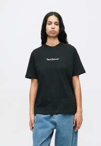 Mulher com cabelo longo e escuro, vestindo uma t-shirt preta da New Balance e jeans azuis, em pé contra um fundo branco simples.