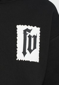 Čierna mikina s výraznou bielou náplasťou so zvlnenými okrajmi, na ktorej je výrazná čierna štýlizovaná grafika "FV".
