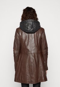 Brun läderjacka med en svart, vadderad huva. Har en figurnära siluett, sömdetaljer och dragkedjeprydnader på ärmarna.