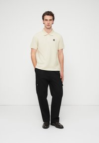 Μπλουζάκι πόλο από βαμβάκι εκρού με κοντά μανίκια, γιακά και μαύρο έμβλημα. Συνδυάζεται με μαύρο παντελόνι τύπου cargo και καφέ παπούτσια.