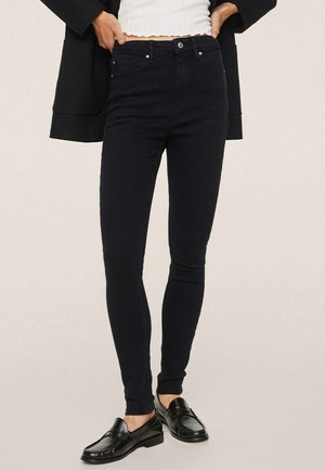Person iført sorte skinny jeans, hvid ribbet kort top, sort blazer og sorte loafers, stående med hænderne på hofterne.