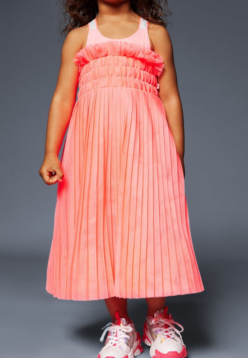 Enfant portant une robe corail sans manches plissée associée à des baskets blanches et roses épaisses, debout devant un fond gris.