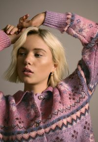 Neulejakku vaaleanpunaisen ja laventelin sävyissä, jossa on geometrinen kuvio, pyöreä kaulus ja kaksi napinläpeä. Ribbaus hihansuiden reunassa tuo tekstuuria.