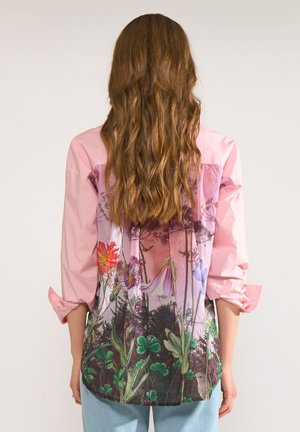 Frau mit langen, welligen Haaren, die ein pinkes Hemd mit bunten Blumen- und Naturmustern auf dem Rücken trägt, kombiniert mit hellblauen Jeans.