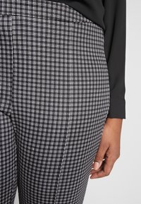 Pantaloni a scacchi in bianco e nero con un design aderente, caratterizzati da un tessuto texturizzato e dettagli visibili della vita. Una mano poggia sul lato.