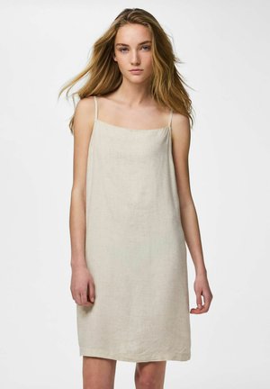 Tânără cu păr lung și blond purtând o rochie fără mâneci, bej deschis, până la genunchi, pe un fundal alb simplu.