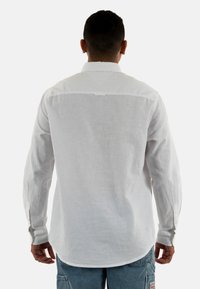 Camicia bianca a maniche lunghe realizzata in tessuto di lino. Presenta un colletto e polsini con bottoni, un orlo curvato e un design con yoke posteriore.
