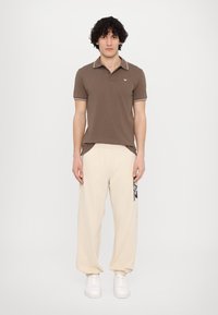 Brown polo shirt with a contrasting collar, paired with cream harem pants featuring a black graphic print. White sneakers complete the look.