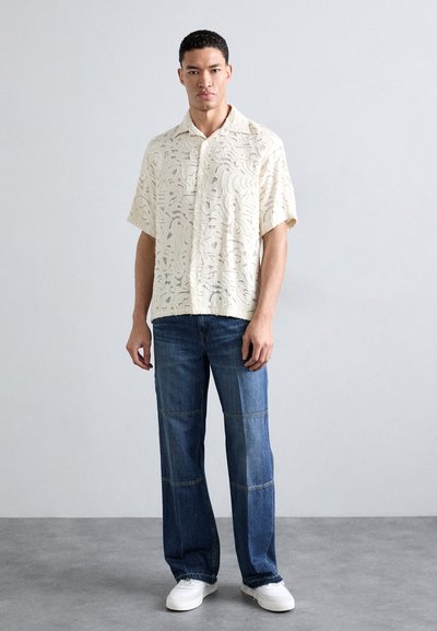 Kortermet, elfenbensfarget mønstret skjorte med en avslappet passform, kombinert med blå patchwork denimbukser og hvite sneakers.