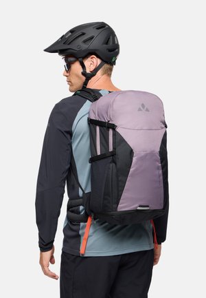 Man draagt een zwarte fietshelm, zonnebril, een shirt met lange mouwen en een grote paars-zwarte rugzak met heup- en borstbanden.