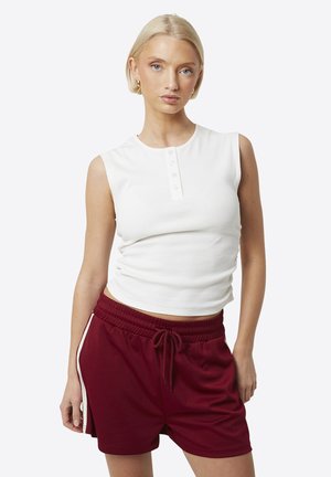 Kobieta o krótkich blond włosach, ubrana w bezrękawnik z białej, zapinanej na guziki bluzki i bordowe szorty z paskiem ściągającym oraz bocznymi paskami, stojąca na tle białego tła.