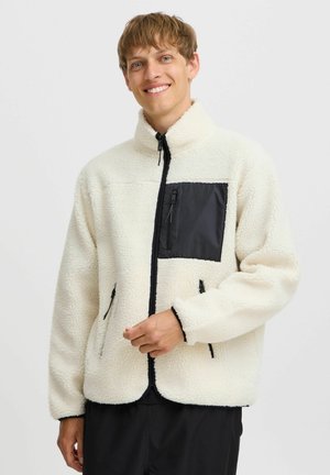 Chaqueta de forro polar blanca con bolsillo en el pecho negro con cremallera, bolsillos laterales, cuello alto y puños elásticos, que presenta un ajuste relajado y una textura suave.