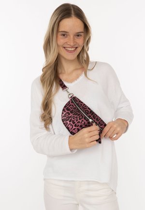 Lächelnde Frau in weißer Kleidung, die eine lilafarbene Umhängetasche mit Leopardenmuster hält und zumacht, vor einem einfarbigen weißen Hintergrund.