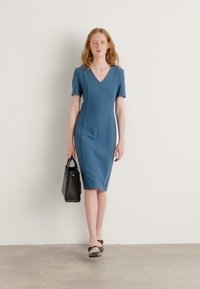 Blue short-sleeve fitted dress with a V-neck, knee-length, featuring subtle vertical seams. Paired with a black handbag and flat shoes.