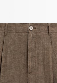 Pantaloni in lino marrone a pieghe con un singolo bottone metallico, taglio dritto e una trama liscia, dotati di tasche laterali.