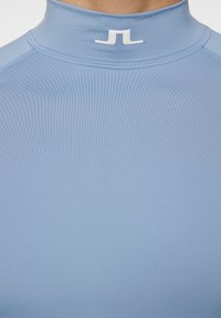 Vaaleansininen korkea kauluspaita, joka on valmistettu sileästä kankaasta. Siinä on pieni valkoinen logo kauluksessa, litteät saumat ja vartalonmyötäinen muotoilu.