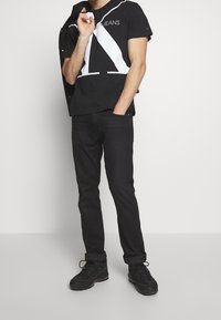 Mies joka pukeutuu mustiin farkkuihin, mustiin lenkkareihin ja mustaan t-paitaan, jossa on valkoinen geometrinen kuvio ja teksti, seisoo yksi käsi taskussa.