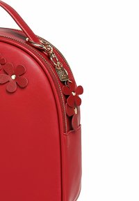 Červená kožená kabelka s kulatým tvarem, zdobená aplikacemi z červené kůže ve tvaru květin a zlatým zipem s květinovým přívěskem na bílém pozadí.
