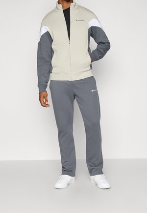 Man draagt een lichtbeige en grijze zip-up Champion jas, grijze joggingbroek en witte sneakers, staand met een hand in de zak.