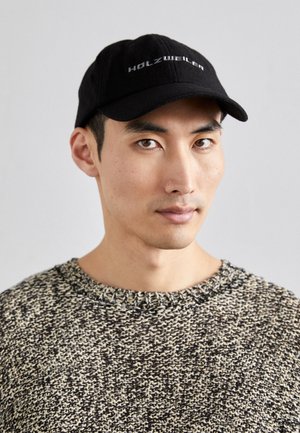 Casquette noire avec le logo brodé "HOLZWEILER" et un visière arrondie, portée avec un pull en maille texturée présentant un motif beige et noir.