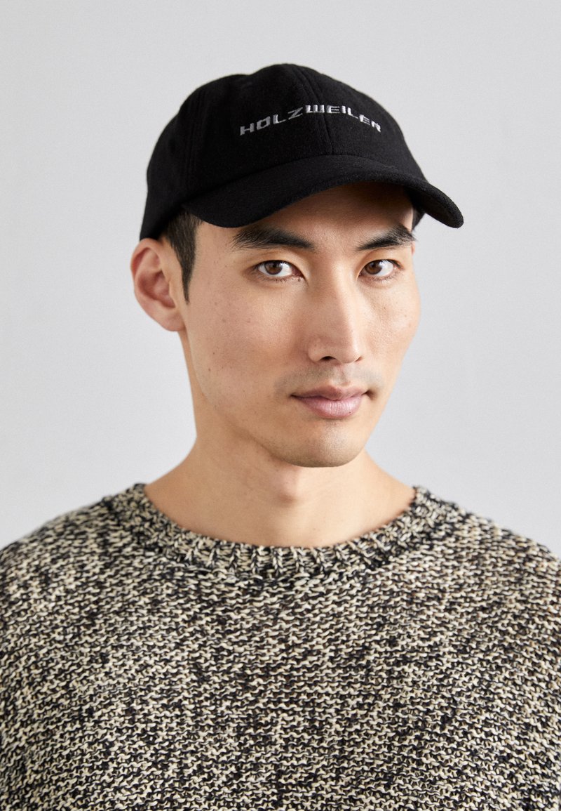 Μαύρο καπέλο με το κεντημένο λογότυπο "HOLZWEILER" και στρογγυλή πινελιά, συνδυασμένο με πλεκτό πουλόβερ με υφή που διαθέτει μπεζ και μαύρο σχέδιο.