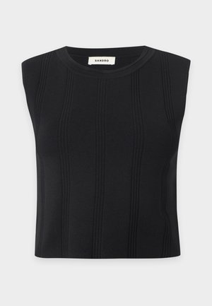 Femme debout portant un crop top noir sans manches, un jean bleu large avec une ceinture marron et des chaussures noires sur un fond blanc uni.