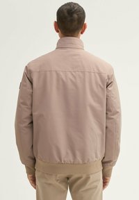 Herenjack in licht taupe, met een hoge kraag, elastische manchetten en een geribbelde zoom. Gemaakt van gladde, duurzame stof met een subtiele glans.