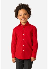 Lächelndes Kind mit lockigem Haar, das ein rotes Langarmhemd mit Knöpfen und dunkle Hosen trägt, steht vor einem einfachen hellen Hintergrund.