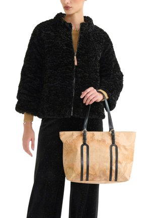 Giacca in pelliccia sintetica nera con colletto alto, abbinata a una borsa tote color cuoio con motivo mappa del mondo e manici in pelle nera. Pantaloni neri.