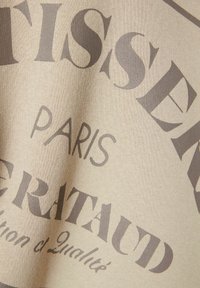 Beige stof met grote, gedurfde grijze tekst waarin de woorden "TISSER" en "PARIS" duidelijk zichtbaar zijn, met een gladde textuur.