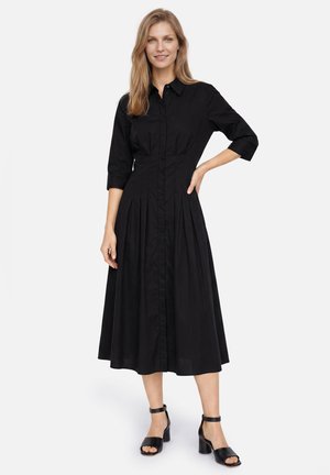 Frau in einem enganliegenden schwarzen Midikleid mit dreiviertel Ärmeln und Knopfleiste vorne, trägt schwarze Sandalen mit knöchelriemen und Blockabsatz.