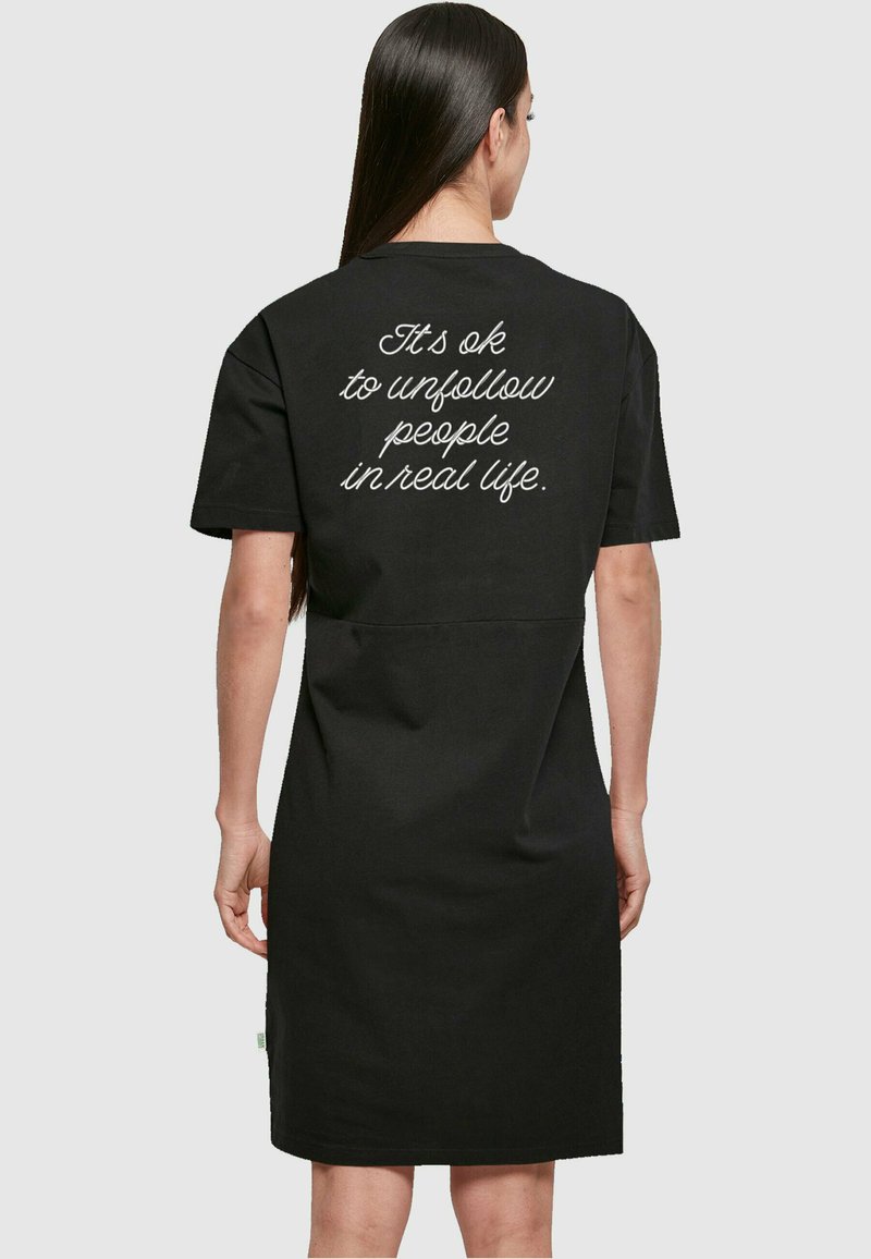 Robe en coton noire à manches courtes, avec un texte blanc dans le dos : « C'est OK de ne plus suivre des gens dans la vie réelle. » Design simple et décontracté.